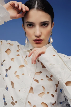 Close-up portrait of a woman wearing a white cut-out shirt and large pearl drop earrings against a solid blue backdrop.