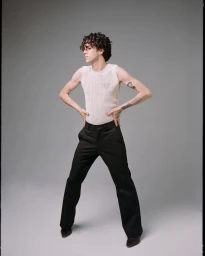 A man wearing a white knit sleeveless top and black trousers standing against a plain grey studio backdrop.