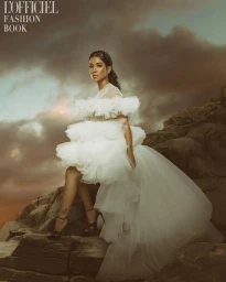 Woman in voluminous white ruffled dress perched on rocky terrain under a dramatic cloudy sky
