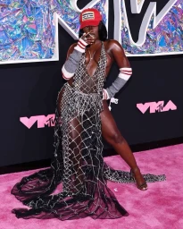 Black woman in sheer crystal mesh gown and red baseball cap on pink carpet with MTV branding