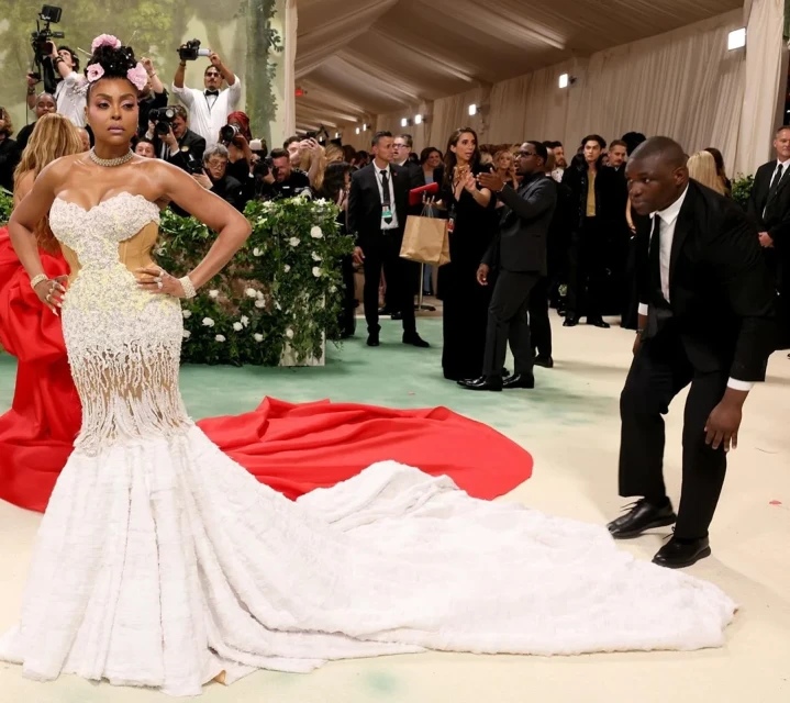 Woman in a white gown posing on a red carpet at an event, surrounded by a crowd and photographers, with a formal indoor setting.