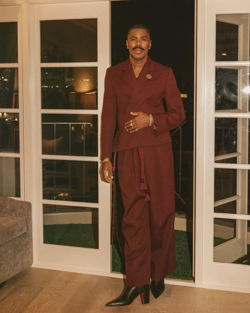 Man in a maroon suit and black heeled boots stands in front of doors leading to a balcony overlooking a city at night.