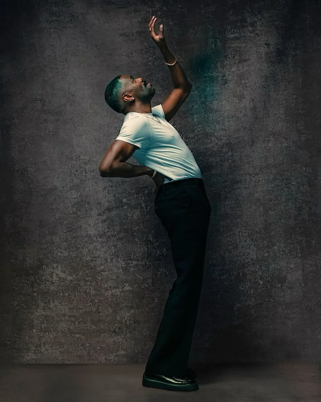 Studio shot of a Black male in white t-shirt and black trousers posing dynamically against a mottled grey background with backlighting.