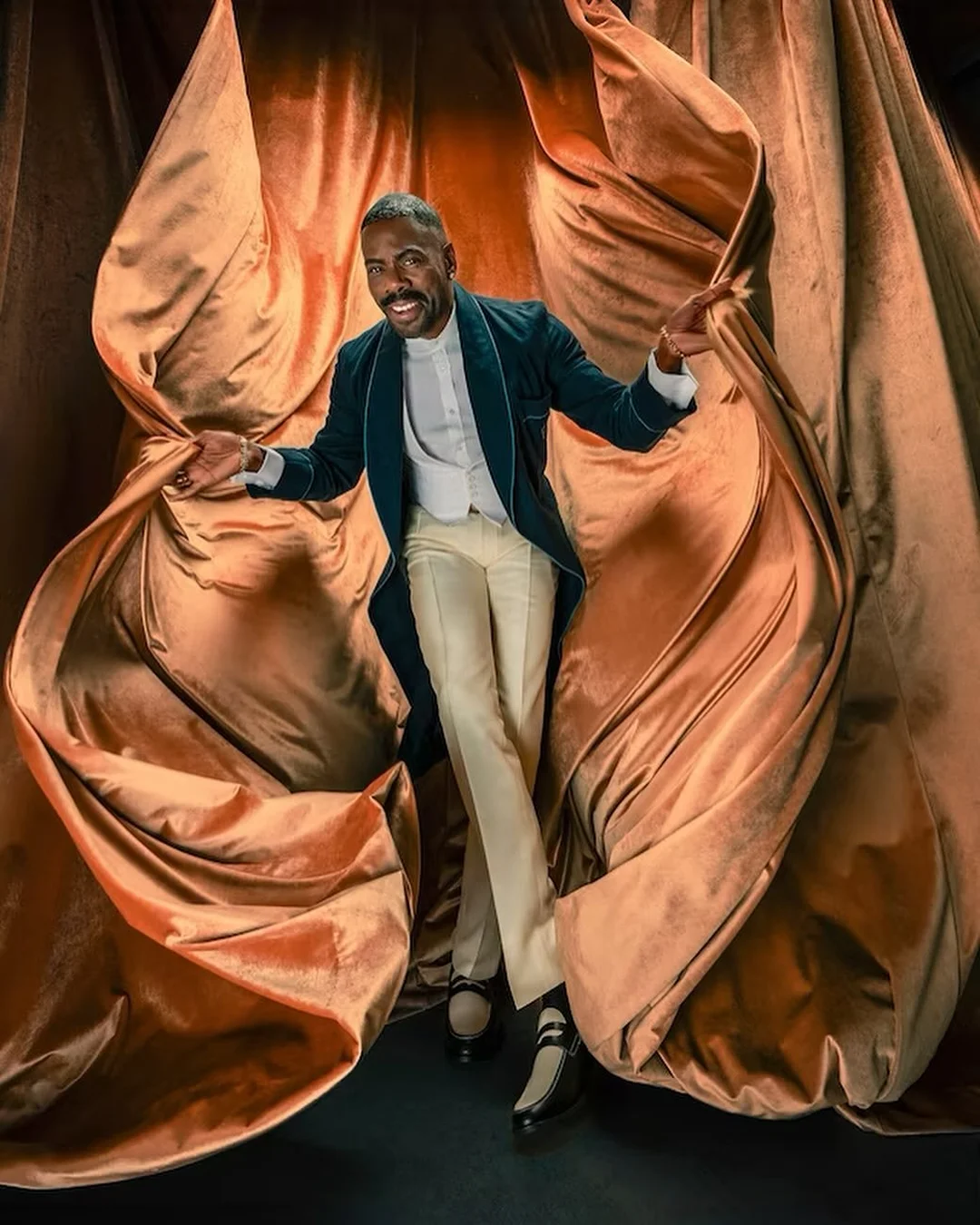 An African American man in a teal blazer and cream pants smiles in front of a draped copper-colored curtain, arms outstretched.