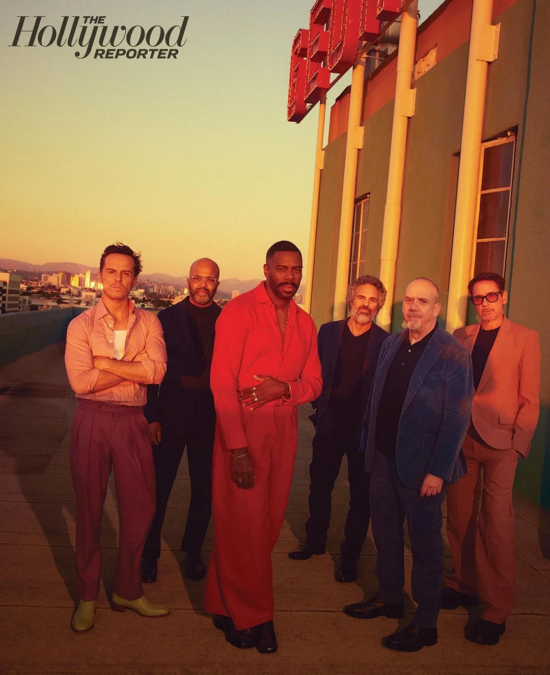 A group of six diverse men stand outdoors in sunlight wearing stylish attire. A building is behind them and a city is in the distance.