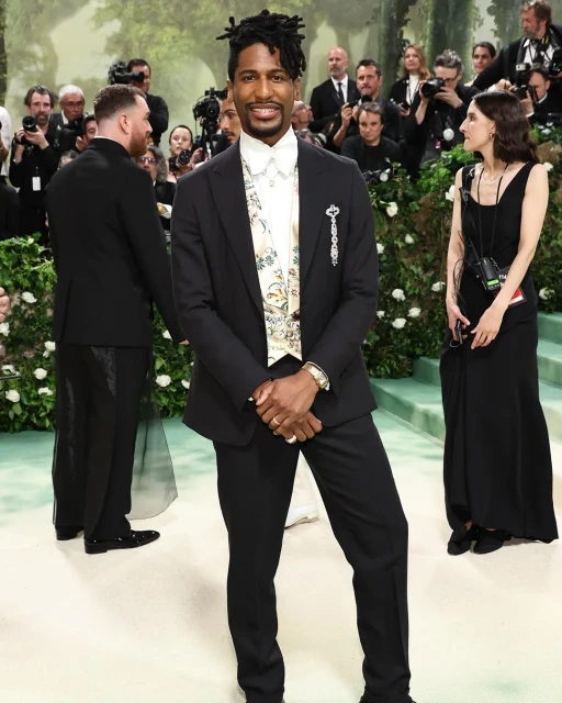 Jon Batiste poses in a dark suit with a floral sash at an event with a press line, greenery and other attendees in the background.