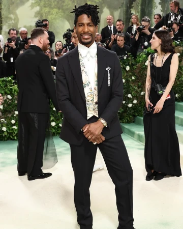 Jon Batiste poses in a dark suit with a floral sash at an event with a press line, greenery and other attendees in the background.