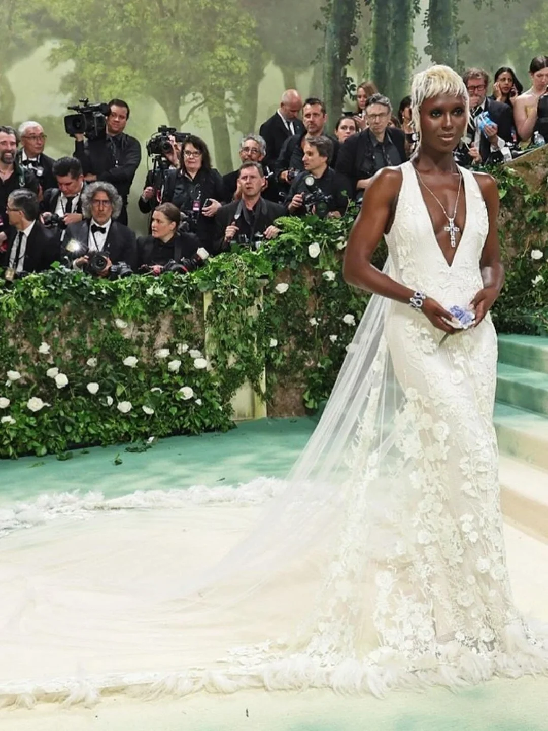 Woman in white lace gown with floral embellishments, fair skin and short light hair, posing on a green carpet at a fashion event.