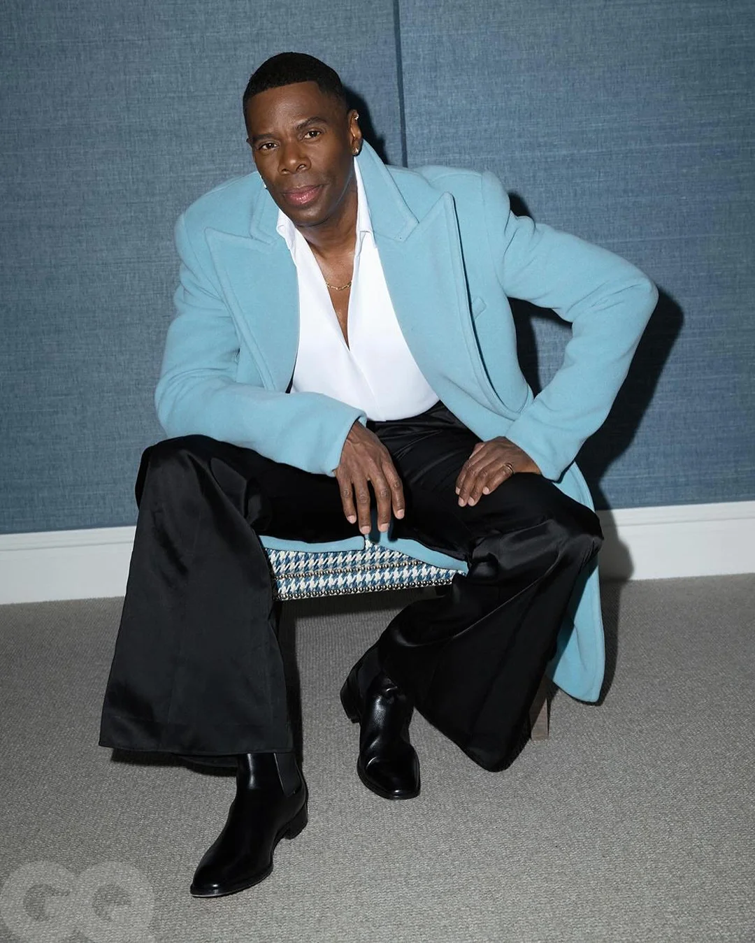 An African-American man sits on a chair in a light blue coat, black pants, and boots, posing against a blue patterned wall indoors.