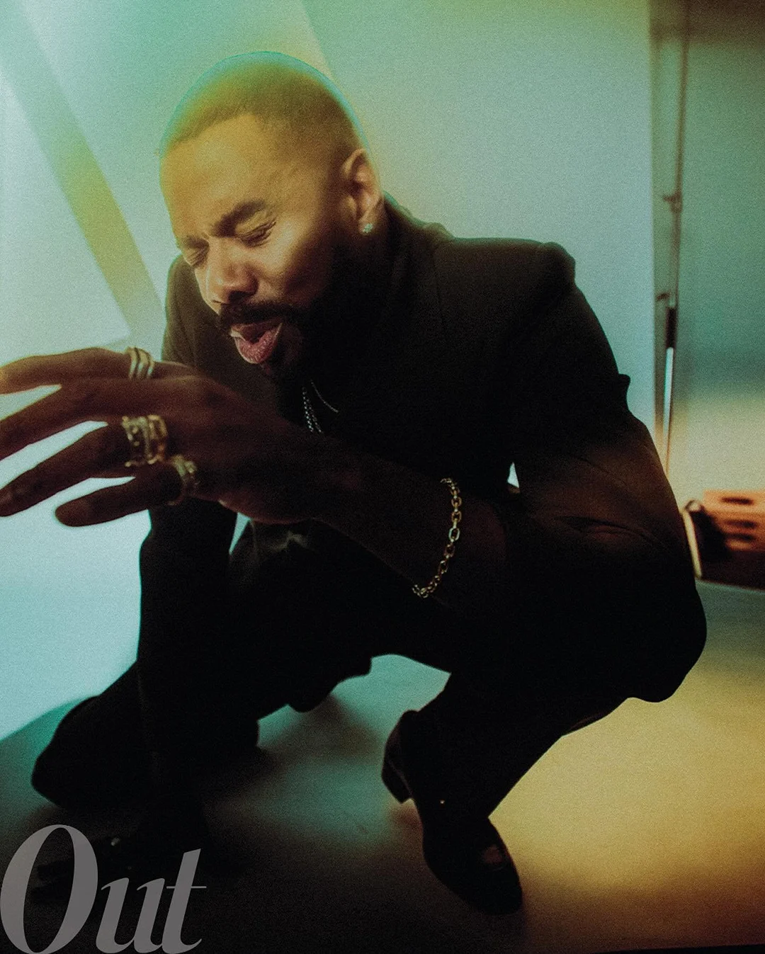 Adult male in black suit poses while crouching; he wears jewelry, with an open mouth and closed eyes in a studio with green lighting.