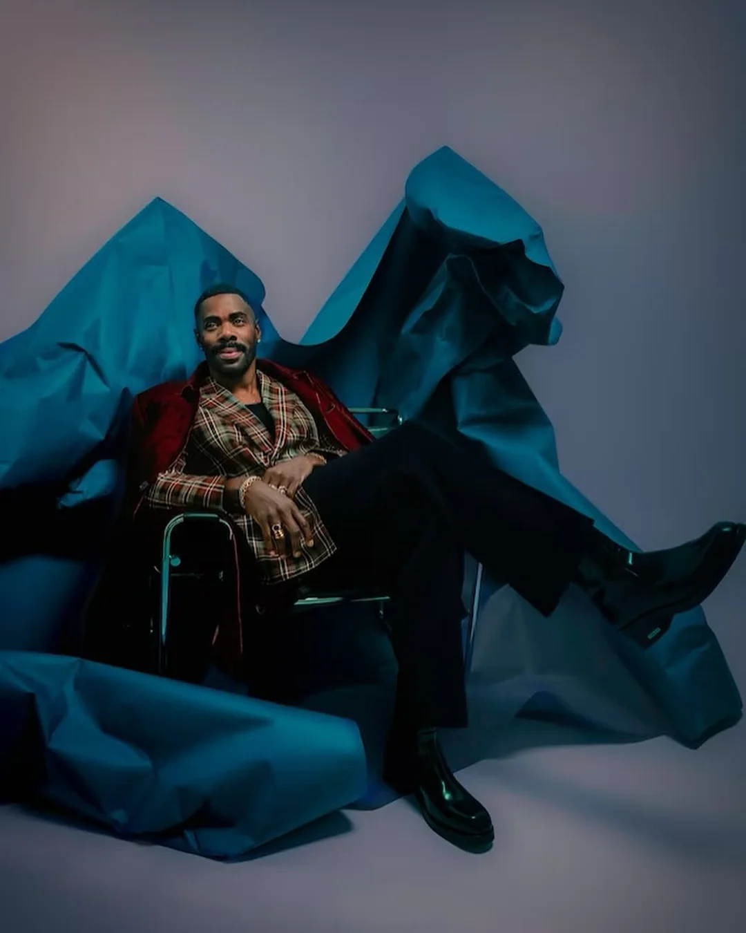 An adult male in a patterned blazer sits in a chair with a modern design, set against a backdrop of draped turquoise fabric.