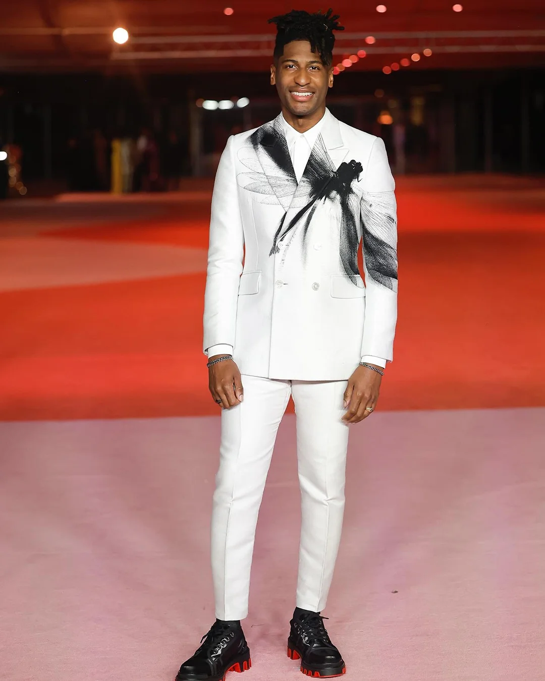 A full shot of a smiling adult male wearing a white suit with a graphic design, posing on a red carpet indoors, with black leather shoes.