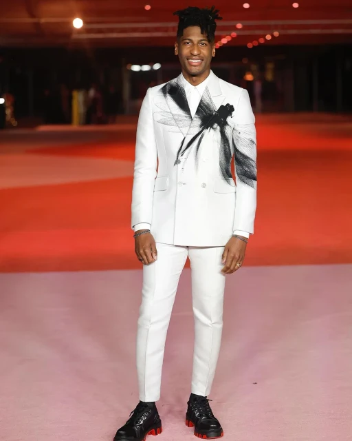 A full shot of a smiling adult male wearing a white suit with a graphic design, posing on a red carpet indoors, with black leather shoes.