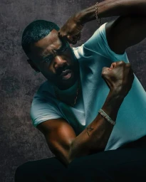 Close-up portrait of a Black man with a mustache wearing a white t-shirt and gold chains, posing with flexed fists.