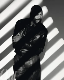 Black and white portrait of a man with a beard wearing a dark jacket and trousers against a wall with striped light and shadow patterns.
