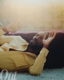 Man wearing a yellow shirt lying down on a shaggy rug with hands raised, styled by Wayman Micah