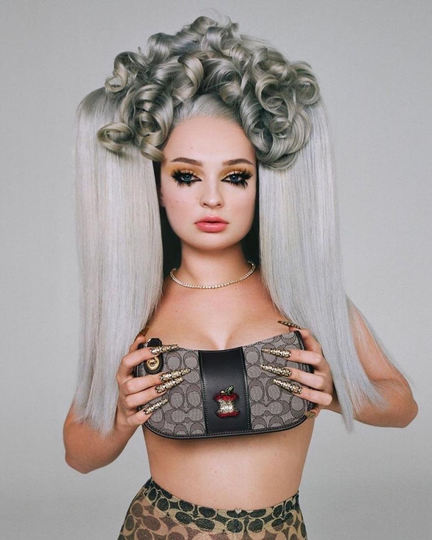 Portrait of an adult woman with gray sculpted hair, heavy eye makeup, holding a Coach purse in front of a gray studio background.