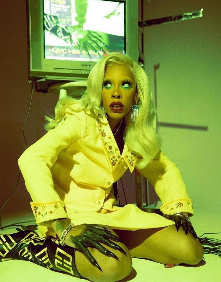 Adult female in yellow suit poses next to vintage television with green lighting, showing long claw-like nails, and blonde wavy wig.