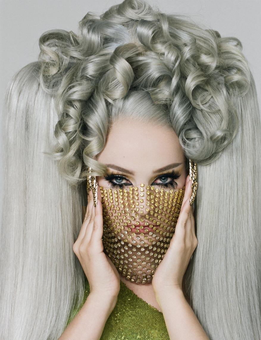 Glamour shot of a woman with voluminous gray hair and a gold mesh face covering, striking a pose in a studio setting.