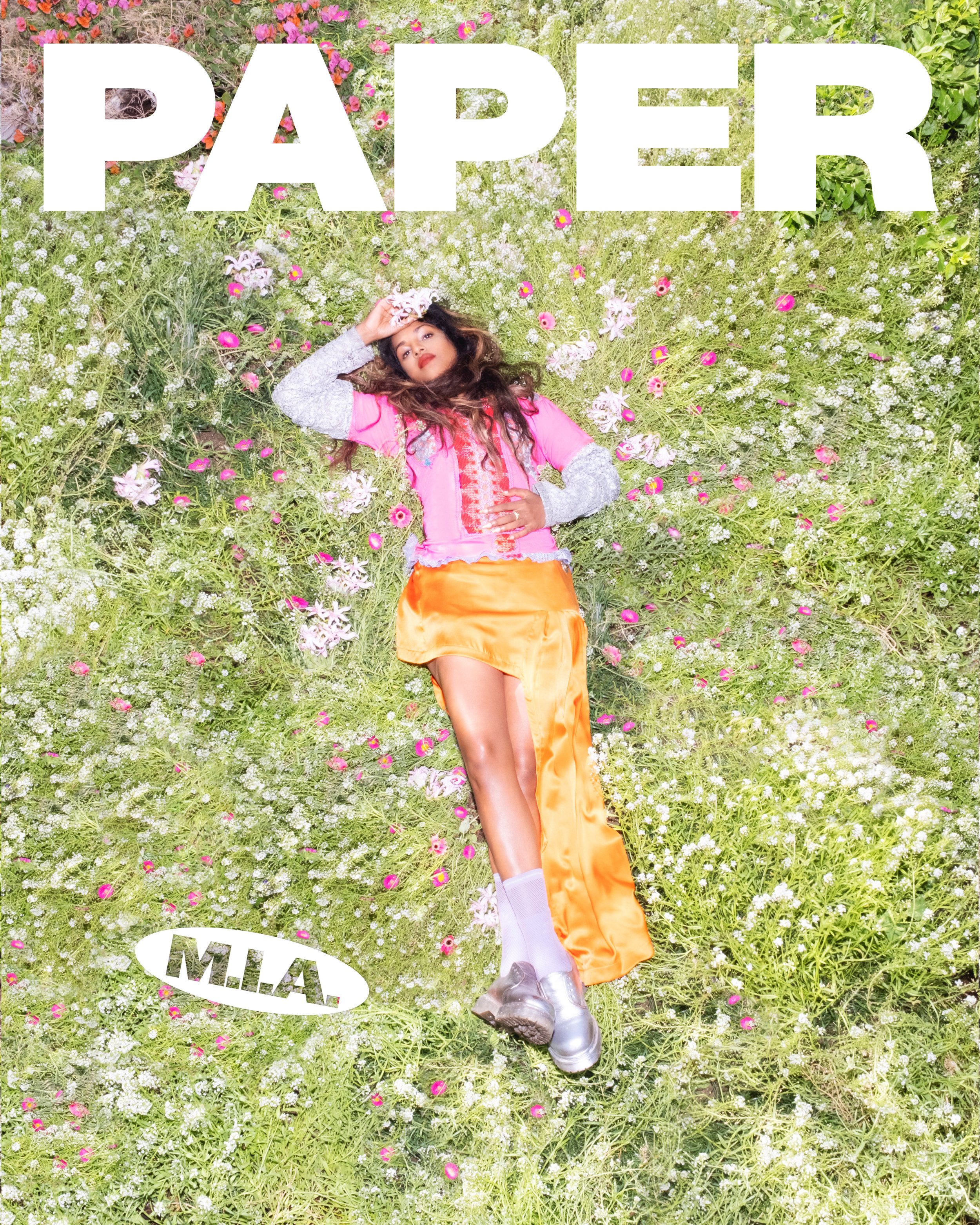 Overhead shot of M.I.A. lying in a field of flowers for Paper magazine cover, wearing a pink top, orange skirt, and silver shoes.