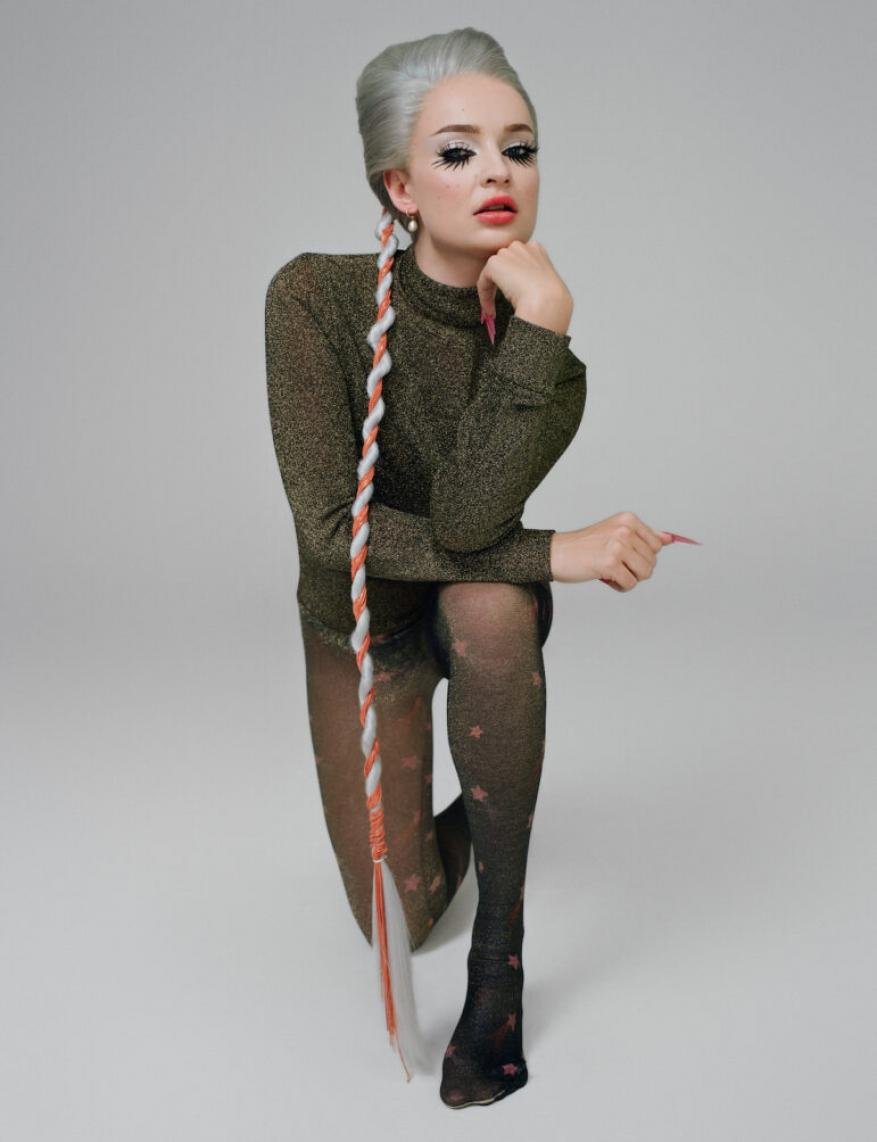 Portrait of a young woman with gray hair in a beehive updo and a long braid, wearing a shiny gold turtleneck and patterned tights, kneeling on a gray surface.
