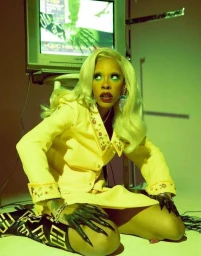 Blonde woman in yellow jacket kneeling on floor with dark painted long nails and television playing in background