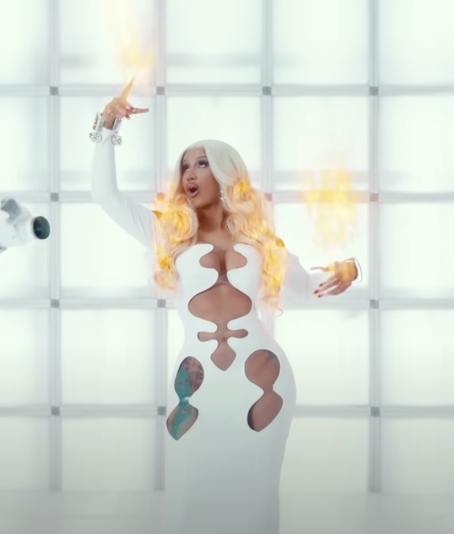 Adult female with blonde curly hair wearing a white gown with cutouts poses with arms raised in a brightly lit, white studio setting.