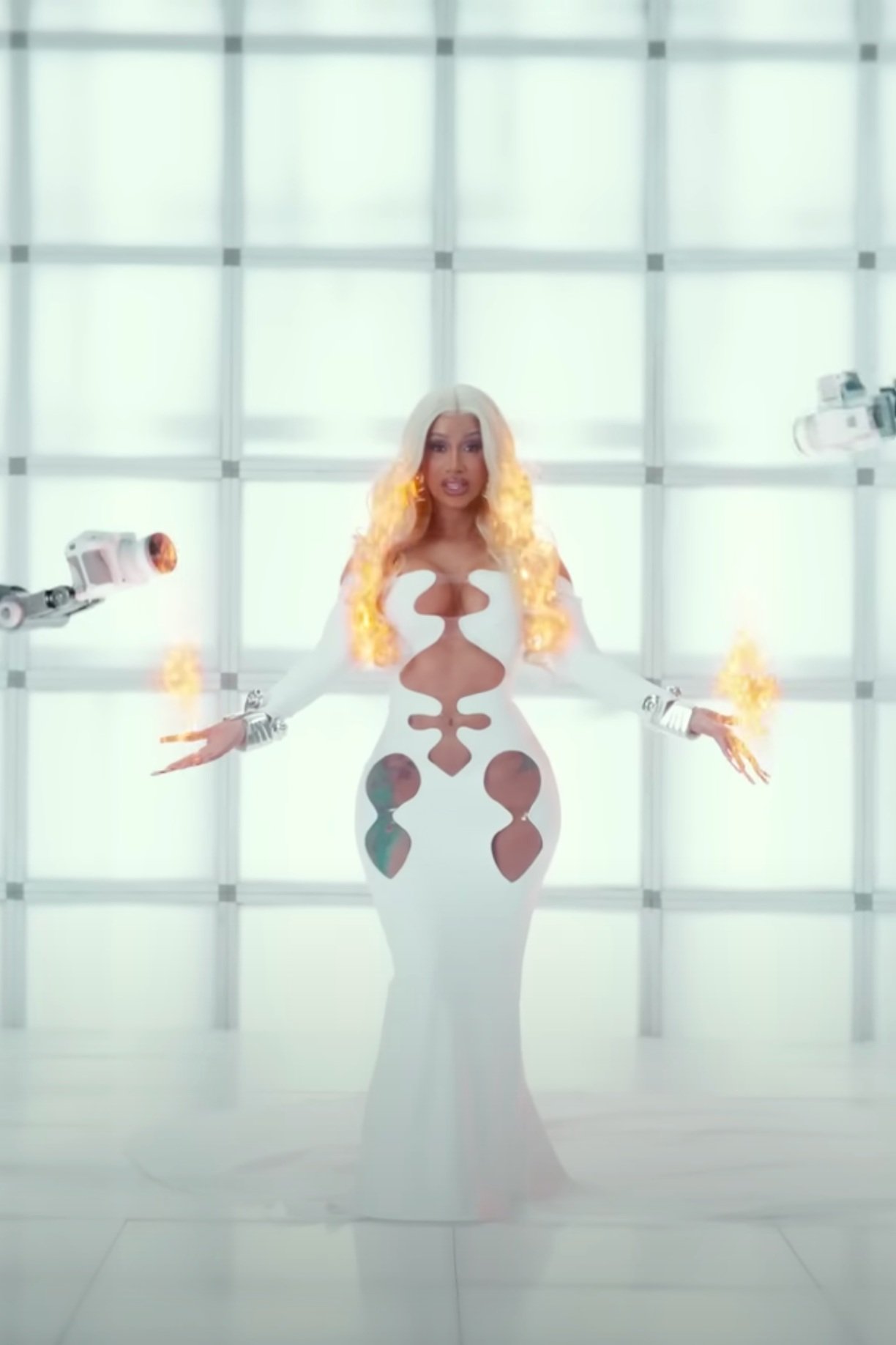Adult female in a white gown, wearing a long blonde wig with flame visual effects, in a brightly lit, futuristic-looking room.