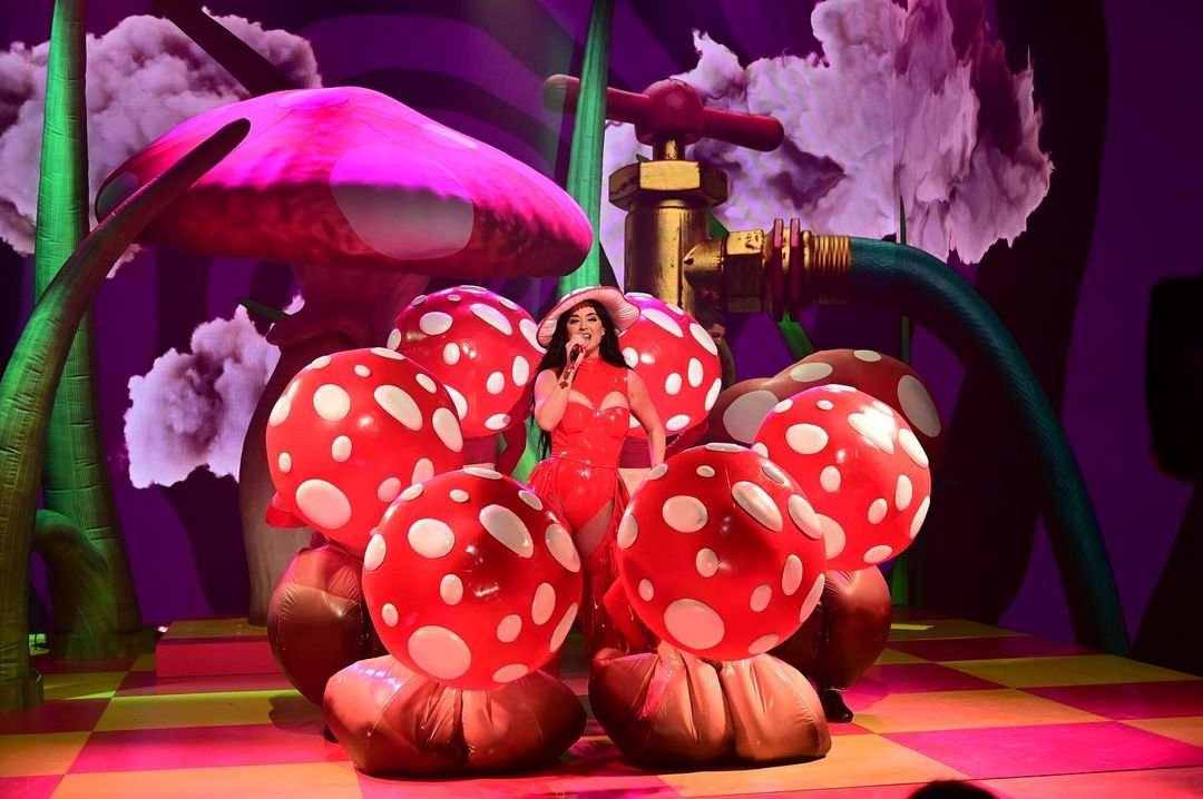 Adult female entertainer in a red latex bodysuit and hat performs on a stage with giant mushroom props and colorful lighting.