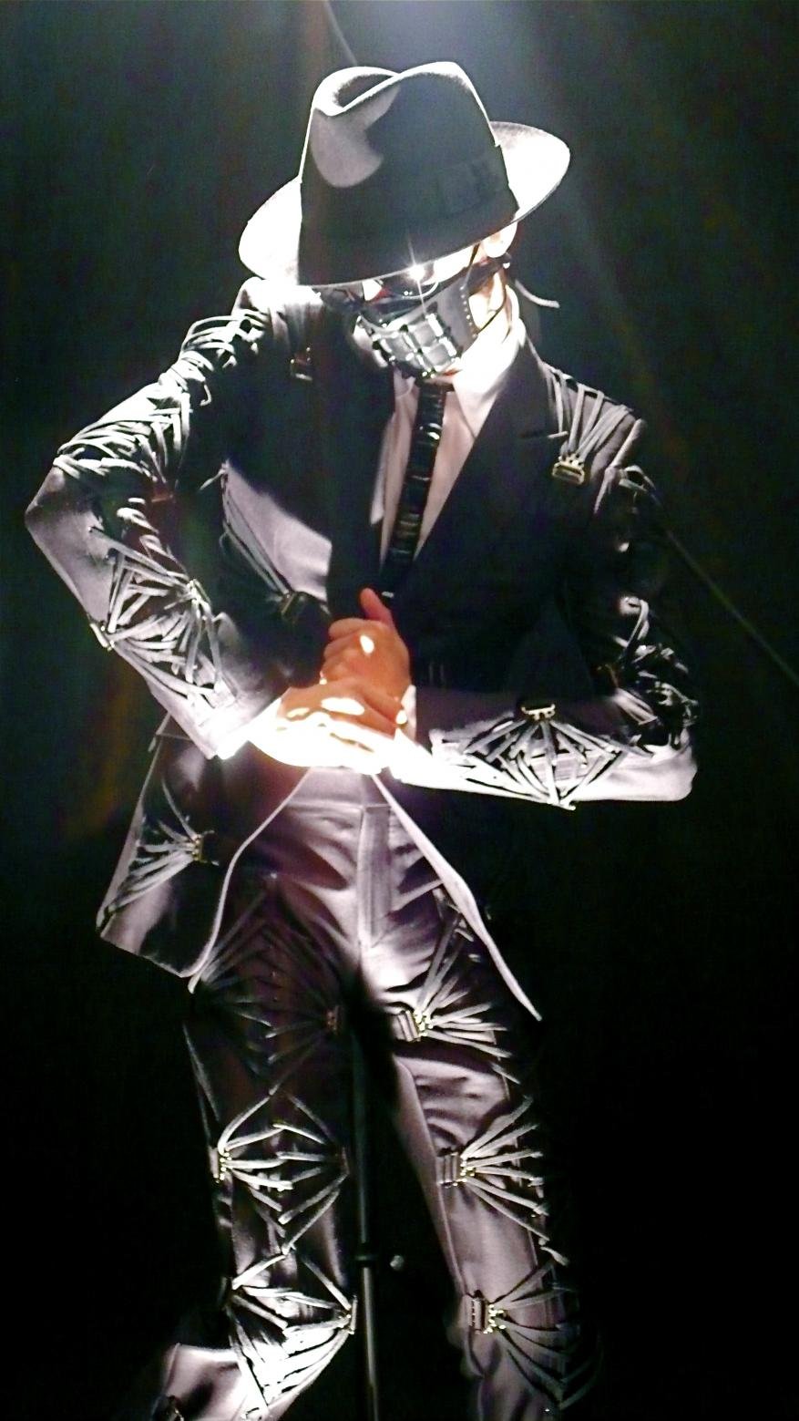 Man in a black suit with geometric patterns and a hat poses under a spotlight, creating a dramatic and stylized presentation.