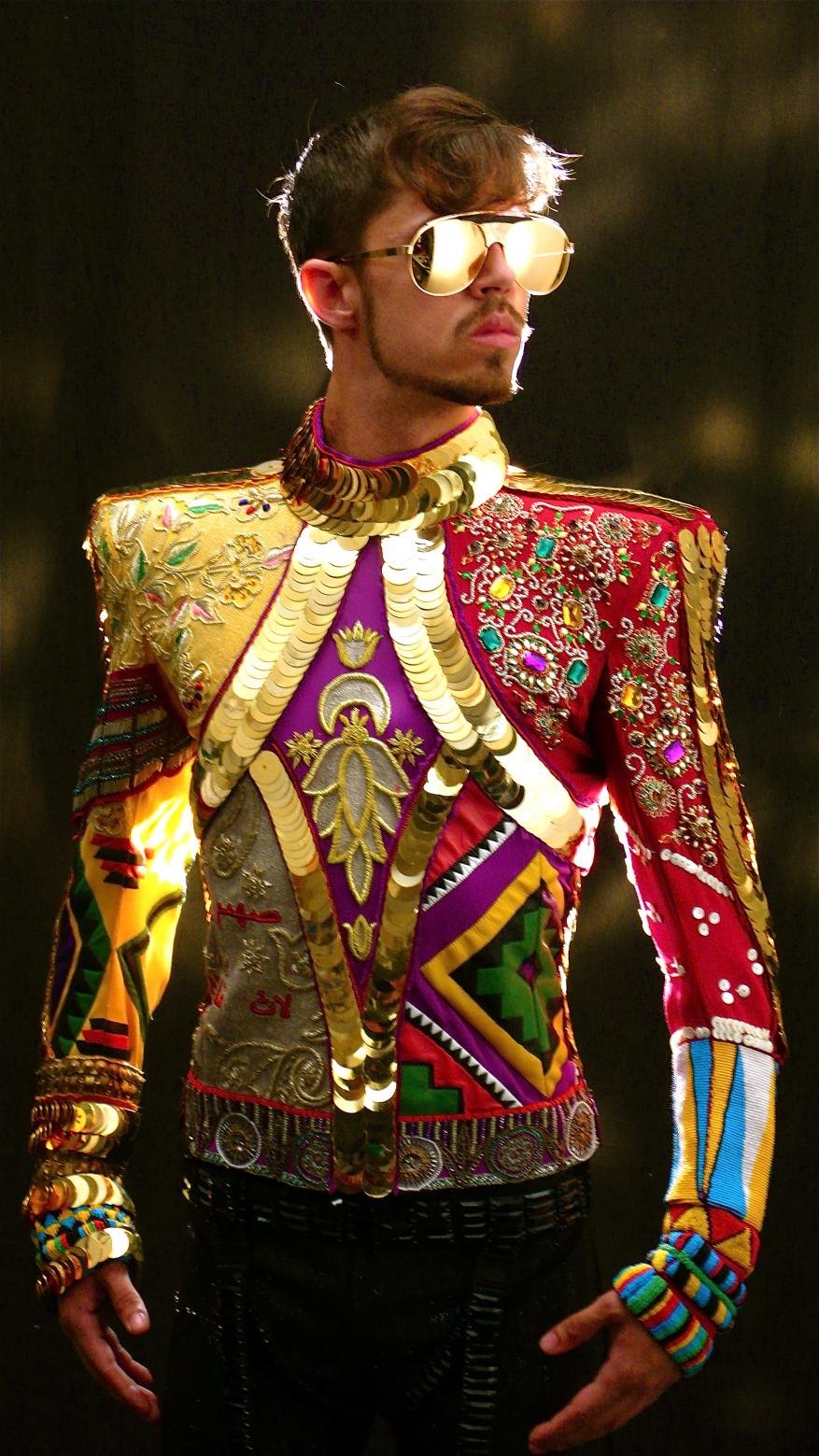 Man in ornate, vintage-inspired jacket, aviator sunglasses, looking left against a dark backdrop. The jacket is colorful, with gold embellishments.