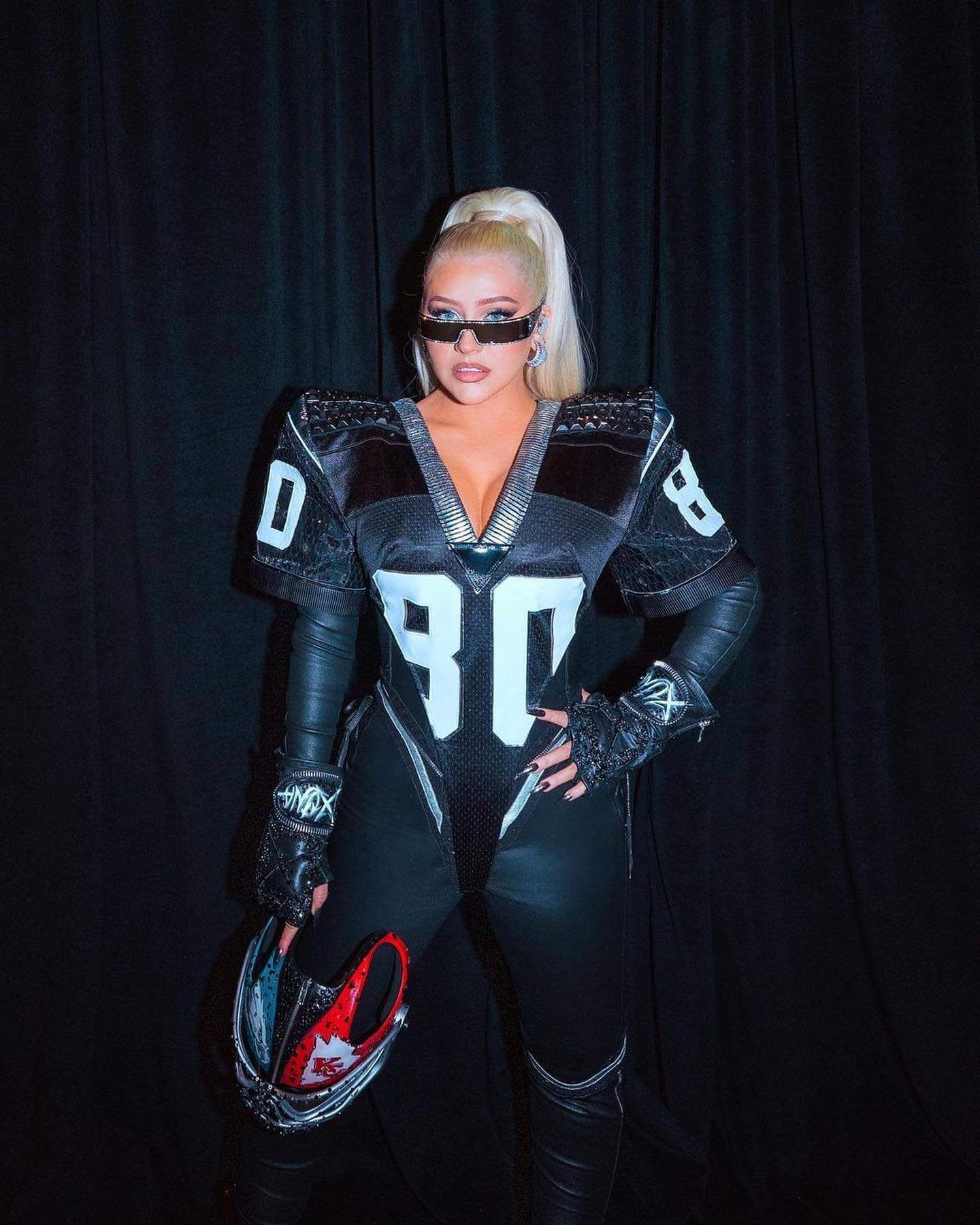Christina Aguilera poses indoors wearing a black American football uniform and sunglasses, holding a helmet with a black curtain backdrop.