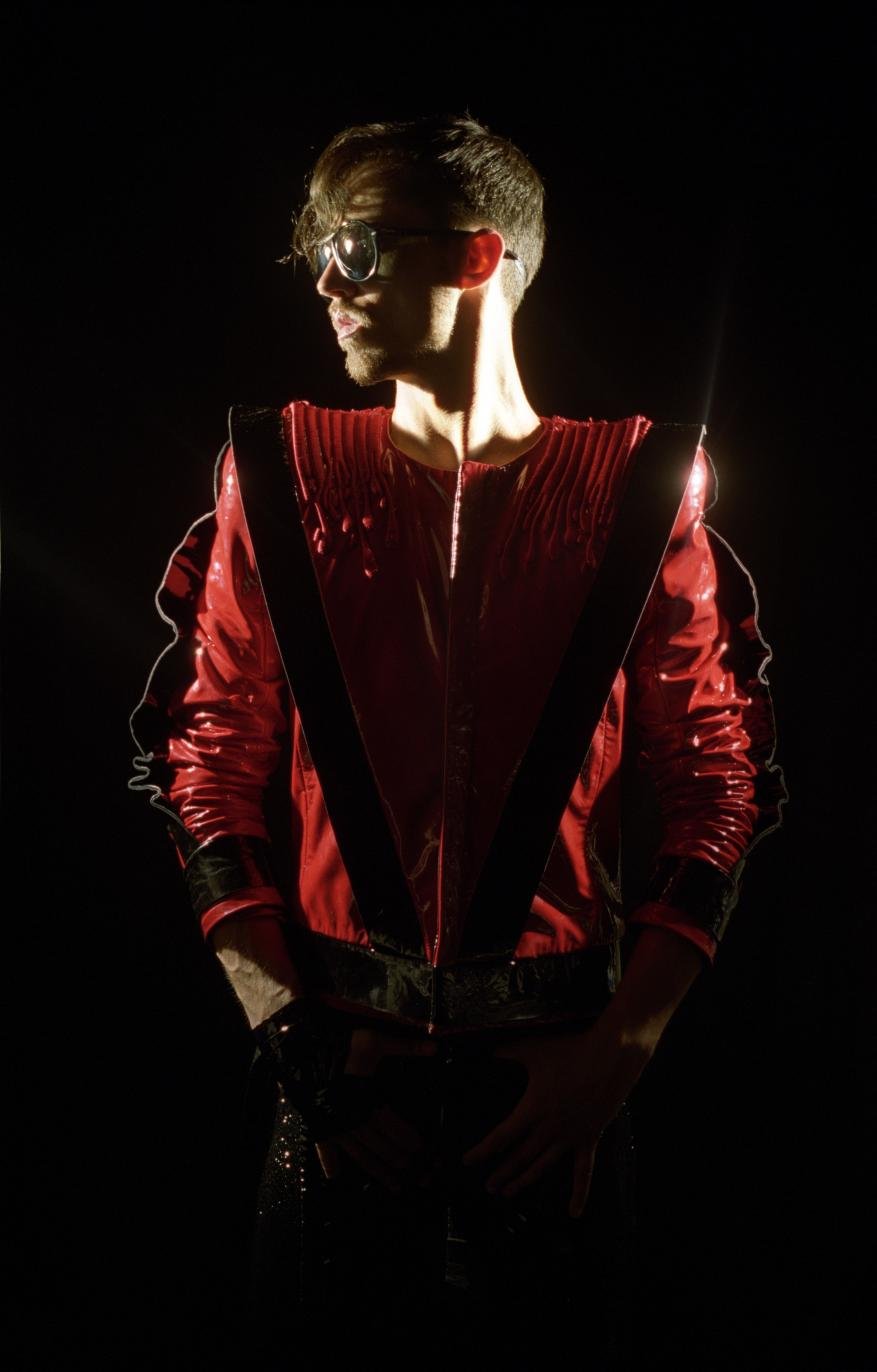 A man wearing glasses and a red jacket with black trim is standing against a dark background, illuminated by a bright light source.
