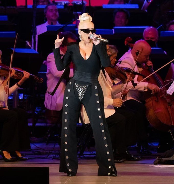 Female singer with microphone performs on stage with symphony orchestra. She wears sunglasses, black pants, and a top.