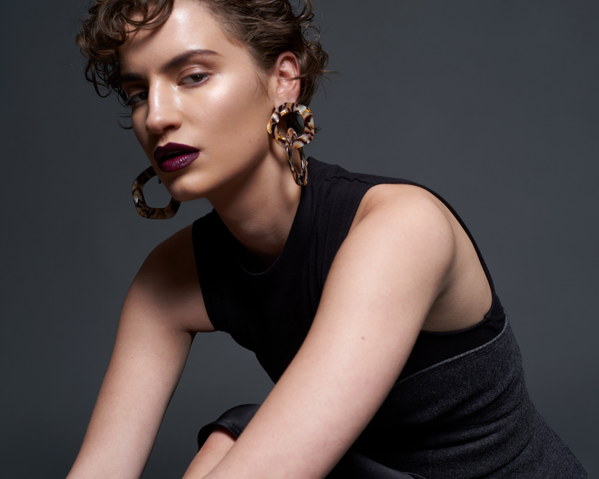 Woman with short curly hair wearing dark lipstick and large tortoiseshell hoop earrings against grey background