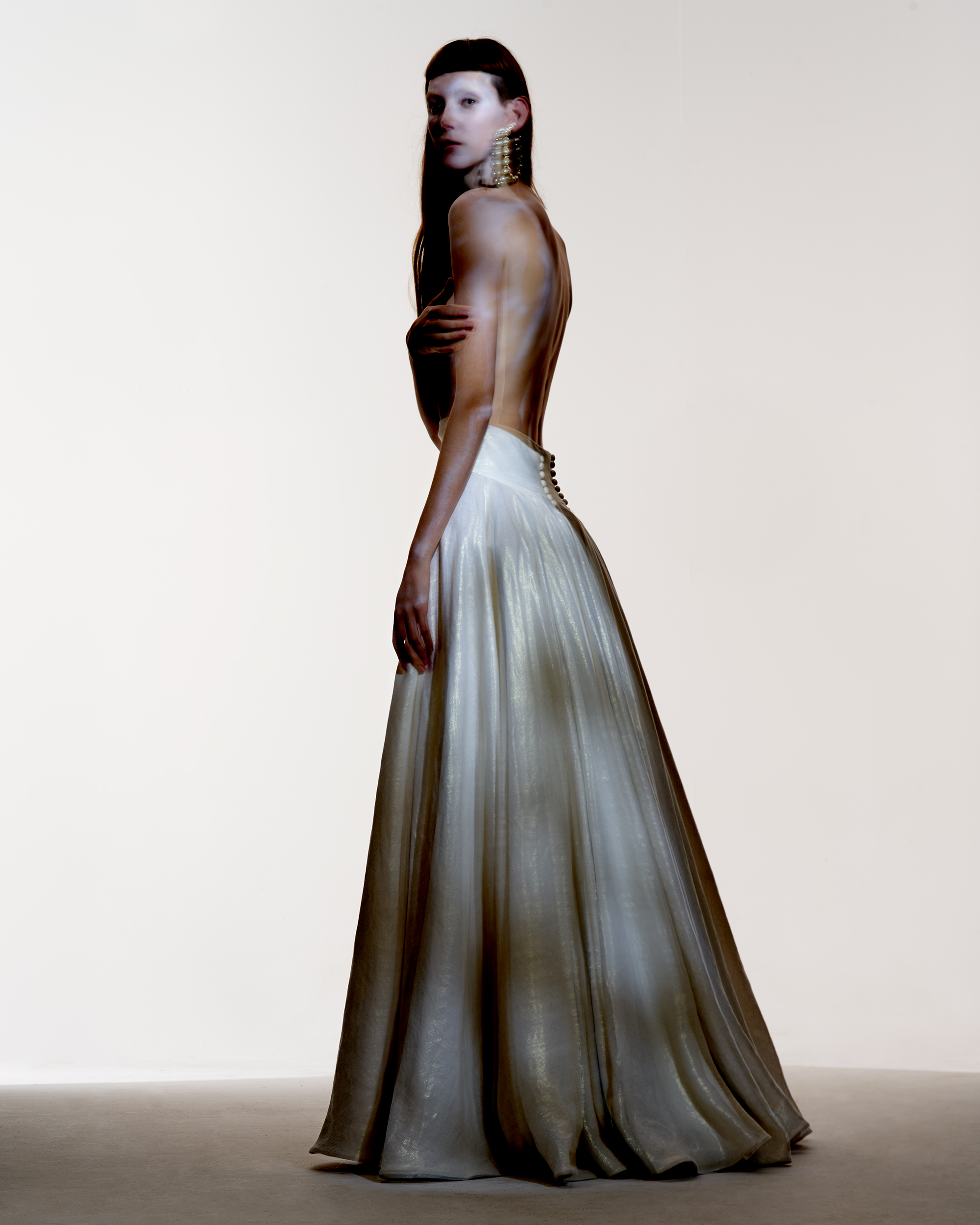 A lone adult female model poses indoors, looking over her shoulder while wearing a low-back, long, flowing white gown and large gold earrings.