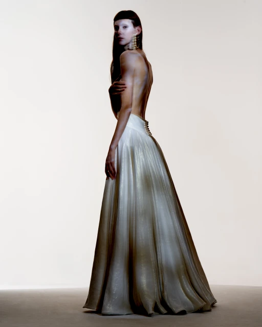 A lone adult female model poses indoors, looking over her shoulder while wearing a low-back, long, flowing white gown and large gold earrings.