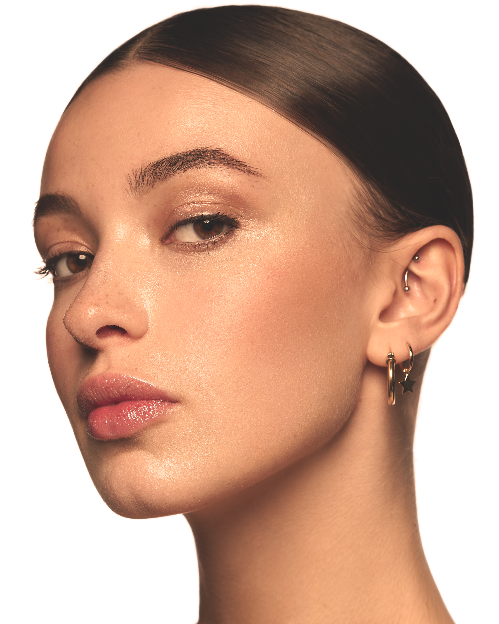 Close-up portrait of a young adult woman with clean, slicked-back dark hair, featuring subtle gold jewelry against a stark white backdrop.