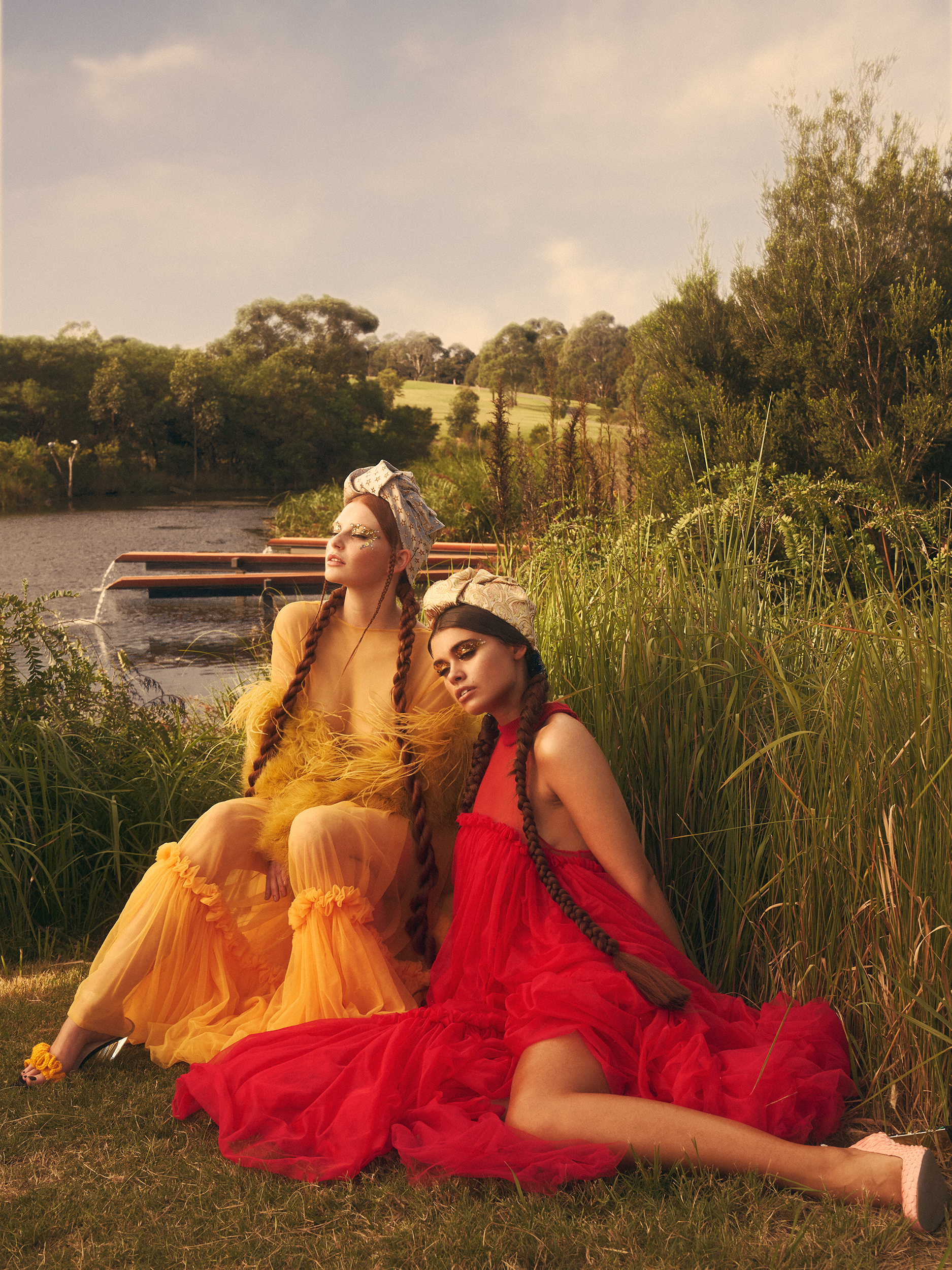 Two women in brightly colored tulle dresses seated outdoors on grass near a lake under a warm sky, photographed by Matt Briggs.