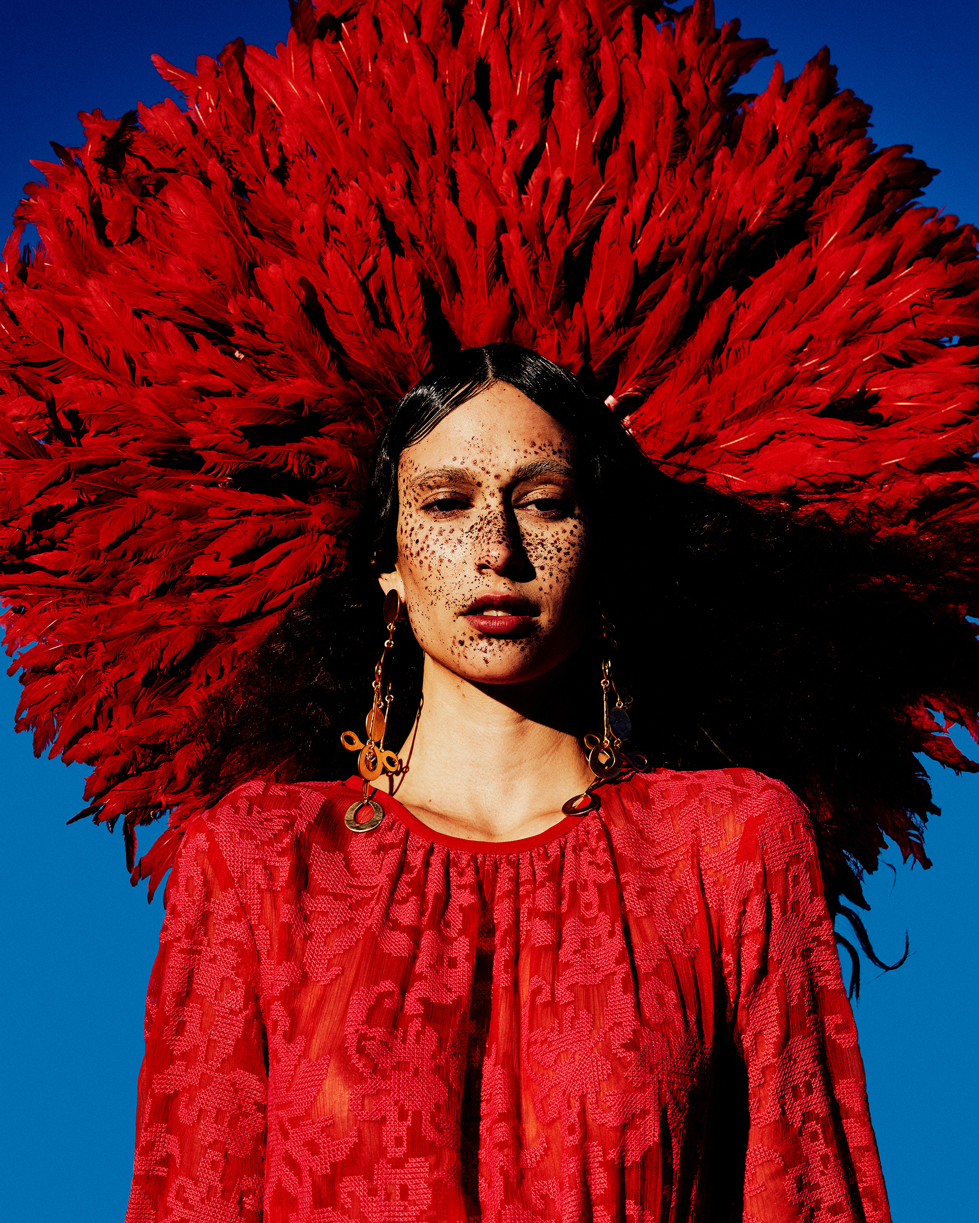 A striking portrait captures a person adorned with a voluminous red feathered headdress, complemented by an ornate red dress against a vivid blue backdrop.