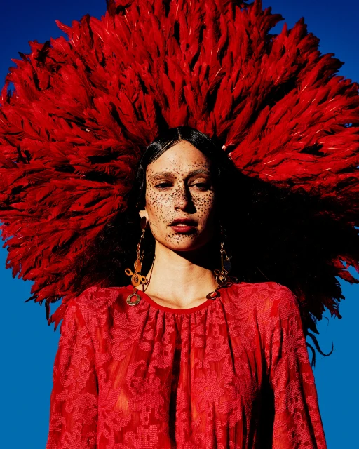 A striking portrait captures a person adorned with a voluminous red feathered headdress, complemented by an ornate red dress against a vivid blue backdrop.