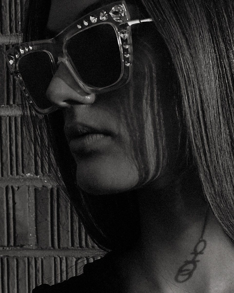 Close-up portrait of an adult woman wearing sunglasses with jewels. Black and white studio shot with a brick wall background.
