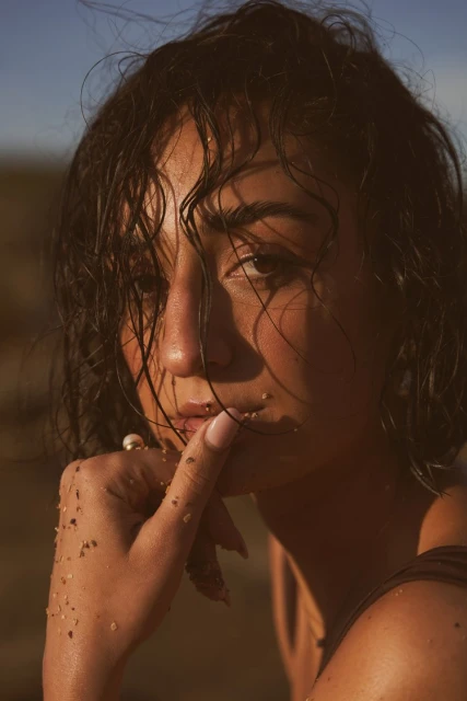 Close-up of a woman with wet, short dark hair in a sun-drenched outdoor setting, her finger to her lips, adorned with sand.