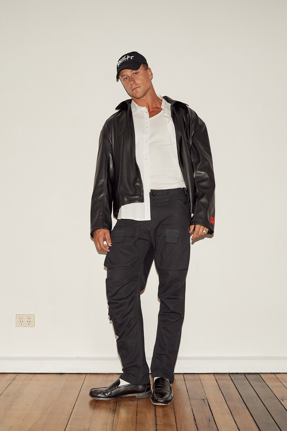 Man wearing a black leather jacket, white shirt, black cap, and dark cargo pants standing against a white wall.