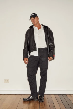 Man wearing a black leather jacket, white shirt, black cap, and dark cargo pants standing against a white wall.