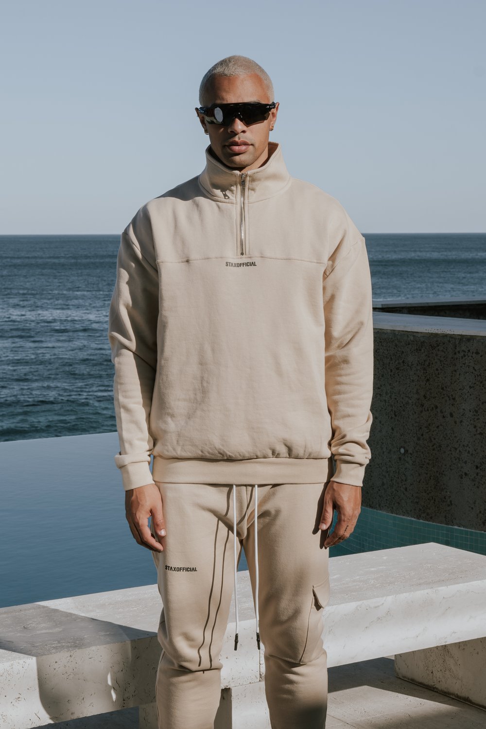 Man with short blonde hair wears sunglasses and a beige tracksuit outdoors in front of the ocean on a sunny day for a fashion shot.
