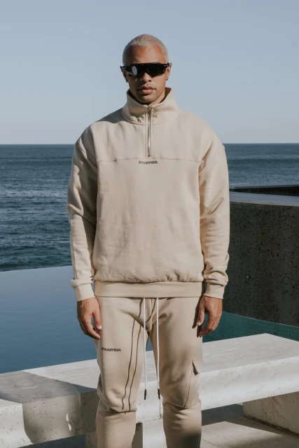 Man with short blonde hair wears sunglasses and a beige tracksuit outdoors in front of the ocean on a sunny day for a fashion shot.