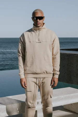 Man with short blonde hair wears sunglasses and a beige tracksuit outdoors in front of the ocean on a sunny day for a fashion shot.