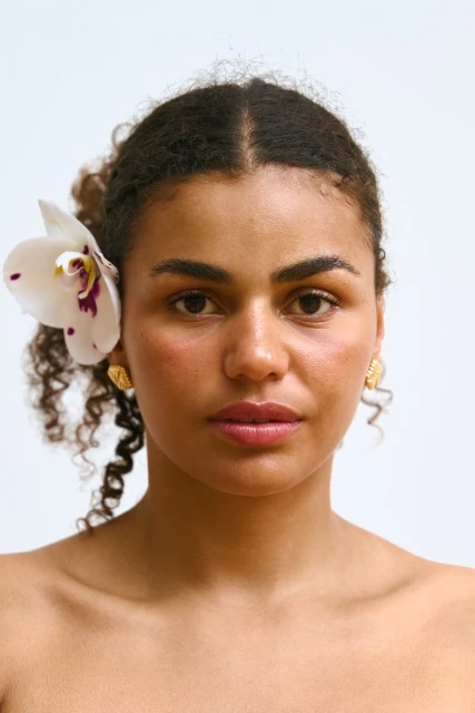 Close-up portrait of an adult woman with curly hair, light skin, and an orchid flower in her hair, posing against a white backdrop.