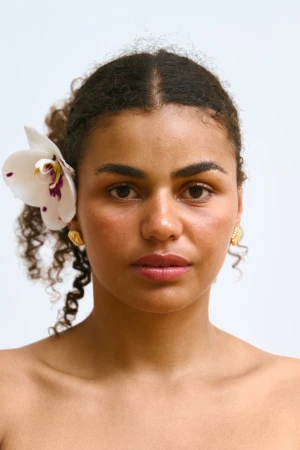 Close-up portrait of an adult woman with curly hair, light skin, and an orchid flower in her hair, posing against a white backdrop.